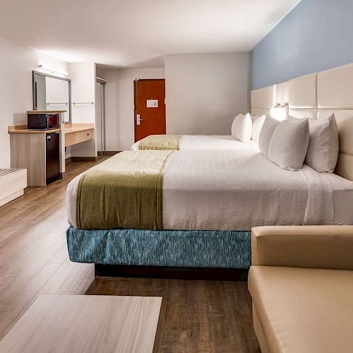 The image shows a hotel room with two double beds, a TV, a microwave, a desk, and a sofa chair on a wooden floor.