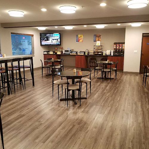 The image shows an empty, clean dining area with wooden floors, tables, and chairs, featuring a buffet area with coffee dispensers and a television.