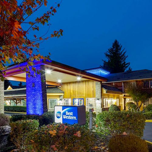 The image shows a Best Western hotel at night, illuminated by blue lights, surrounded by autumn trees and shrubs, with a clear, deep blue sky.