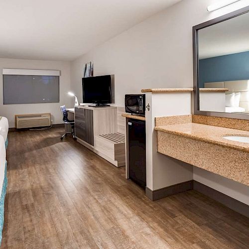 The image shows a modern hotel room with two beds, a desk, a TV on a cabinet, a microwave, a mini-fridge, and a sink with a mirror.