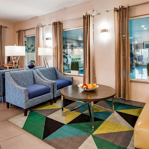 A modern lounge area with blue armchairs, a round coffee table with fruit, geometric rug, and large windows with beige curtains ending the sentence.