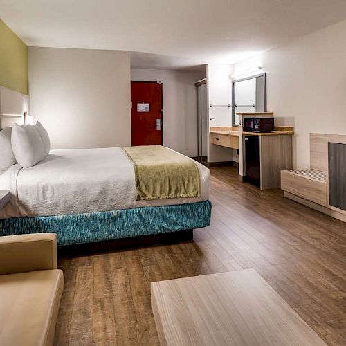 A modern hotel room with a bed, sofa, TV, desk, and a red door in the background. The walls and floor are neutral-toned.