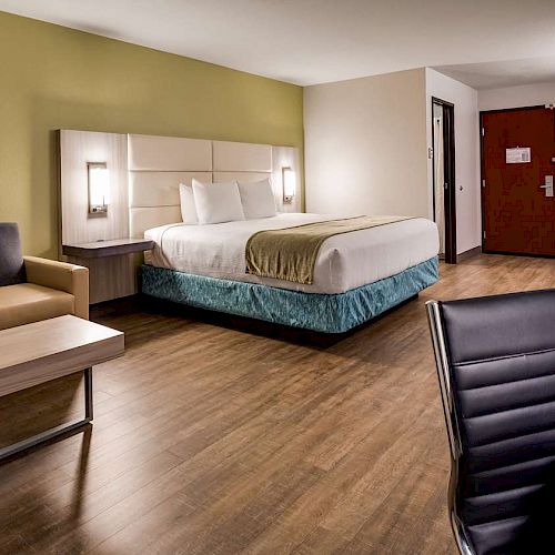 A spacious hotel room with a king-sized bed, sofa, desk, TV, and wood flooring. Modern decor with neutral tones and a red door in the background.