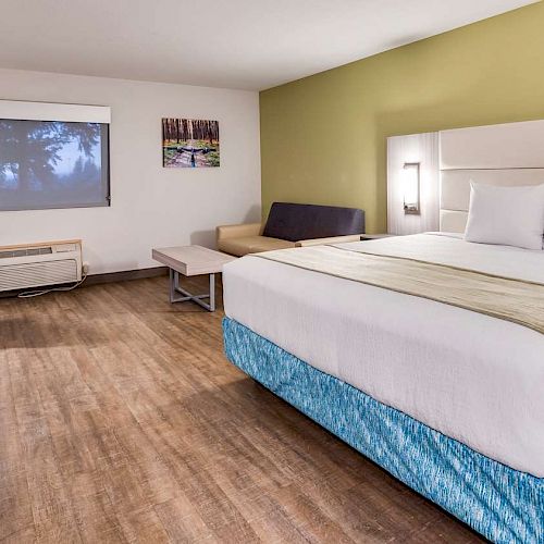 A modern hotel room with a king-sized bed, TV, desk chair, sofa, coffee table, and window. The room has wood flooring and green accent wall.