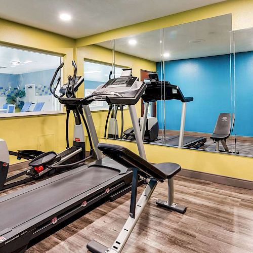 A small gym room with cardio equipment including a treadmill, exercise bike, weights, mirror, wall-mounted TV, and a blue and yellow color scheme.