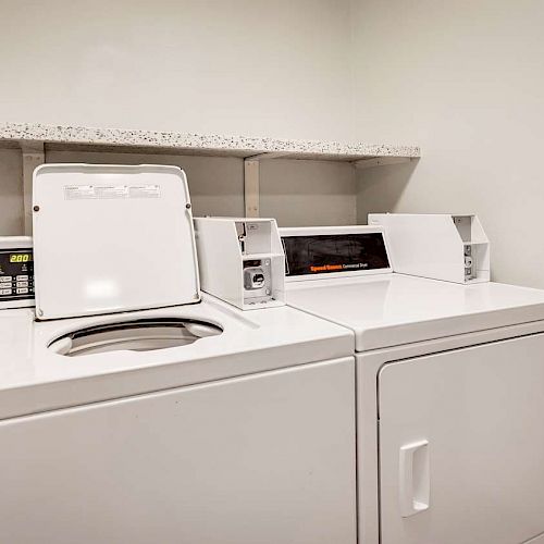 This image shows a small laundry area with a white washing machine and dryer placed side by side under a shelf.