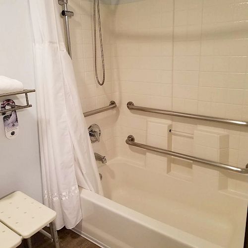 The image shows an accessible bathroom with a shower, equipped with grab bars, a white shower curtain, a wall-mounted showerhead, folding seats, and towels.