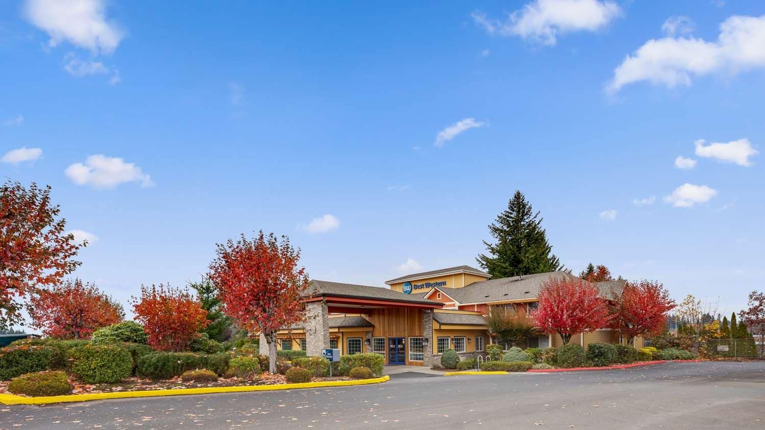 The image shows a building with a sign reading "Best Western" surrounded by landscaped greenery and trees with autumn foliage under a blue sky.