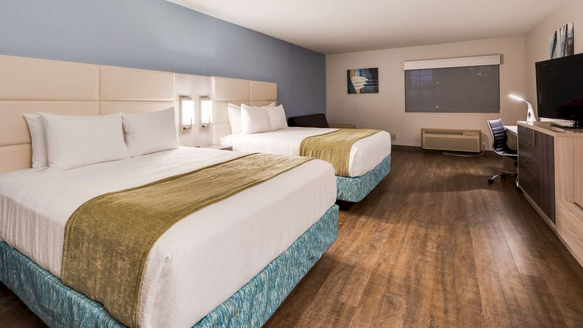 This image shows a hotel room with two large beds, wooden flooring, a desk with a chair, a TV, a window with blinds, and minimalistic decor.