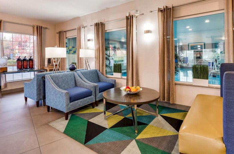 A cozy lounge area with blue chairs, a yellow bench, a round coffee table, and a colorful rug, with large windows and modern decor.