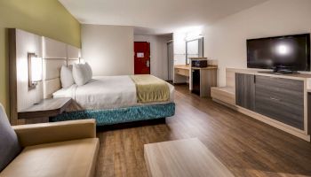 A modern hotel room with a bed, couch, flat-screen TV, desk, and mirror. The room has wooden flooring and a red door in the background.