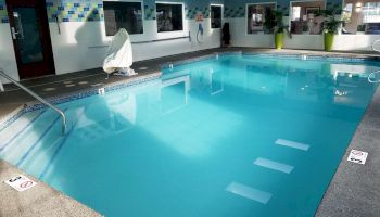 The image shows an indoor swimming pool with clear blue water, surrounded by poolside chairs, windows, and green plants in a spacious room.