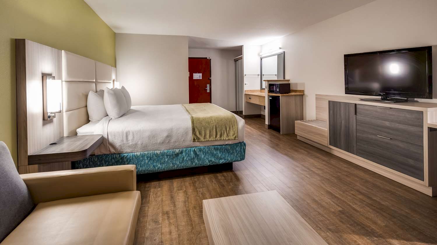 The image shows a modern hotel room with a bed, couch, TV, desk, and a small refrigerator, with wooden flooring and a red door in the background.