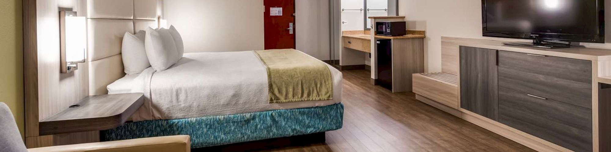 The image shows a modern hotel room with a bed, couch, TV, desk, and a small refrigerator, with wooden flooring and a red door in the background.