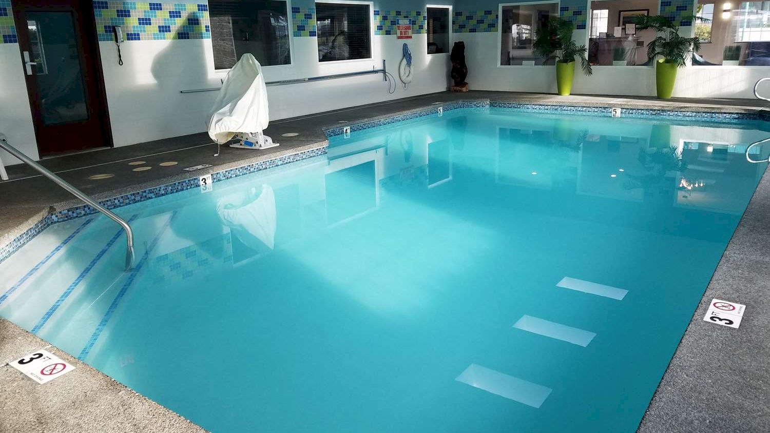This image shows an indoor swimming pool with clear blue water, surrounded by white walls and potted plants. There is a chair lift for accessibility.