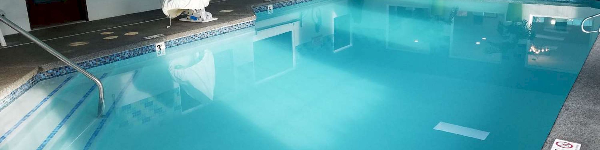 This image shows an indoor swimming pool with clear blue water, surrounded by white walls and potted plants. There is a chair lift for accessibility.