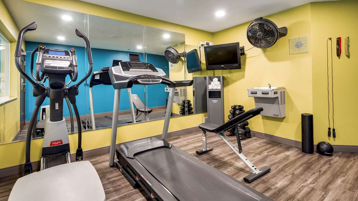 This image shows a small gym with exercise equipment, including a treadmill, elliptical machine, bench, weights, and a television mounted on the wall.