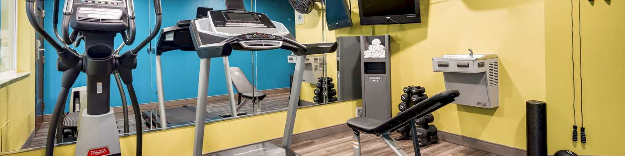 This image shows a small gym with exercise equipment, including a treadmill, elliptical machine, bench, weights, and a television mounted on the wall.