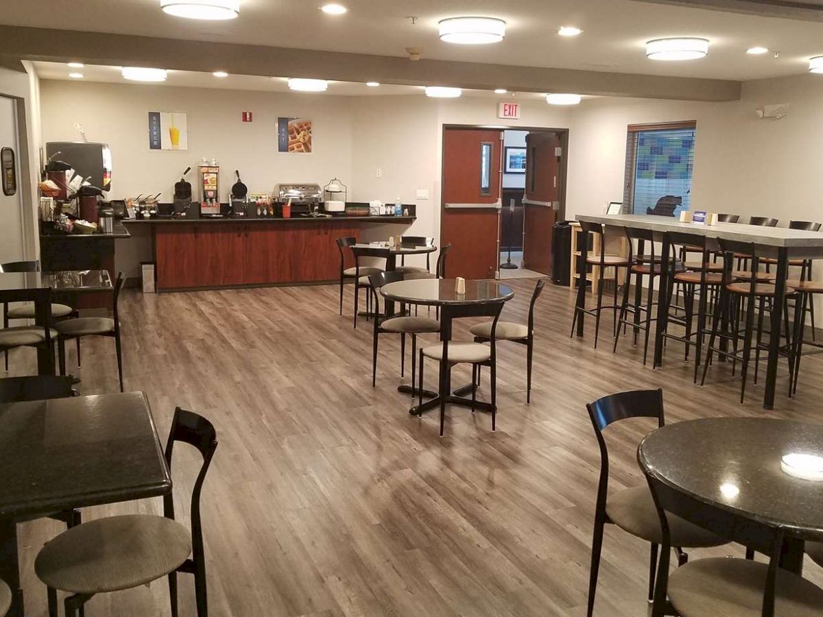 The image shows a clean, well-lit dining area with several tables and chairs, a serving counter in the background, and a person working in the kitchen.