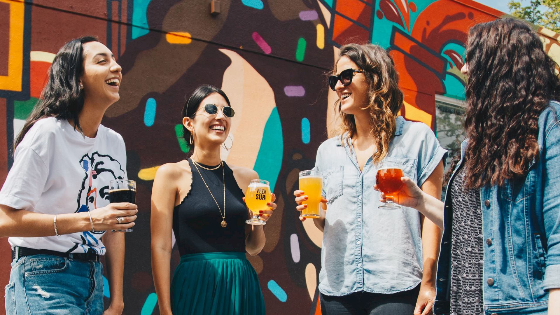 Four women are standing and laughing while holding drinks in front of a colorful mural, enjoying their time together.